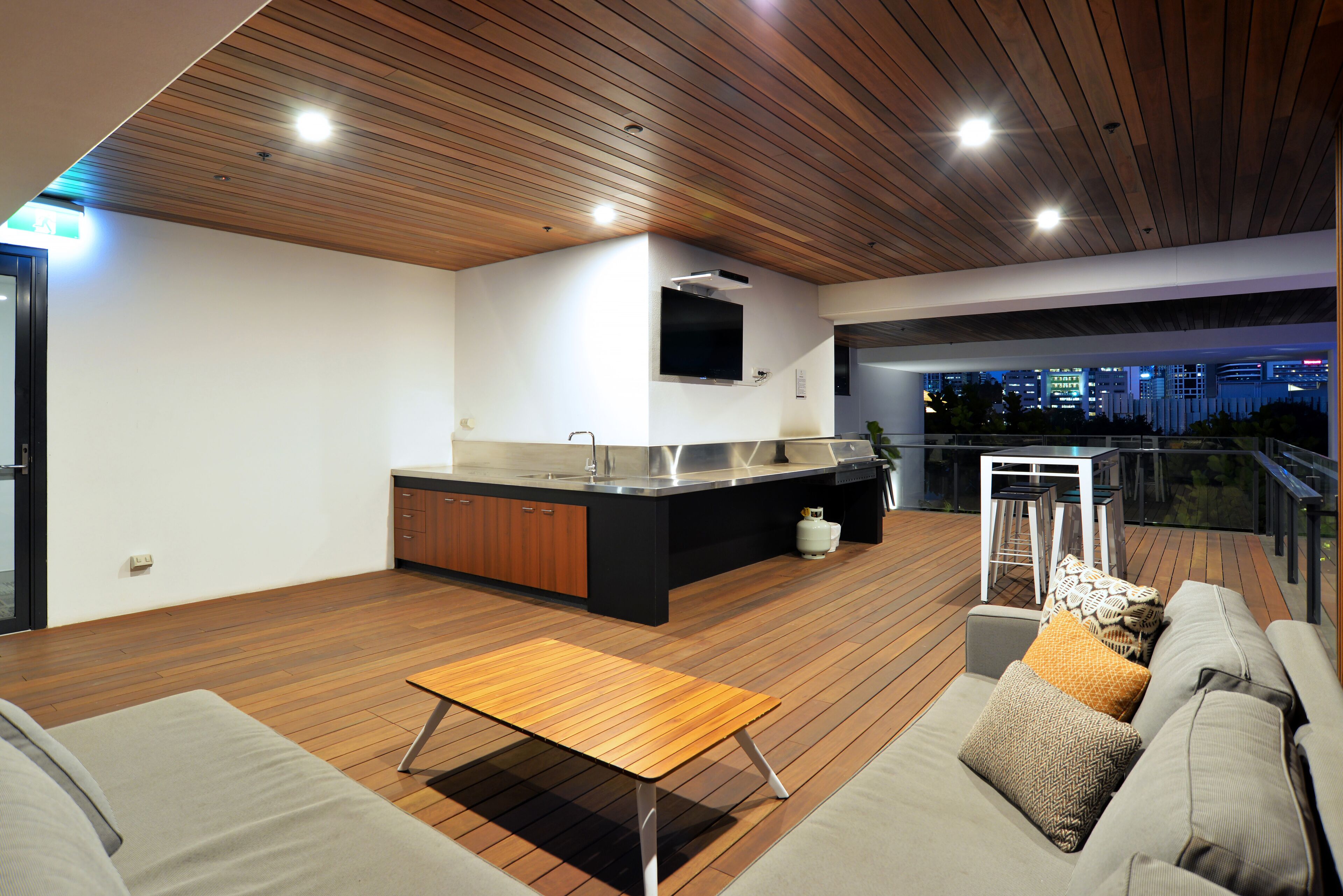Un espacio moderno para disfrutar del aire libre con terraza de madera, cocina integrada, sofá y vistas nocturnas de la ciudad.
