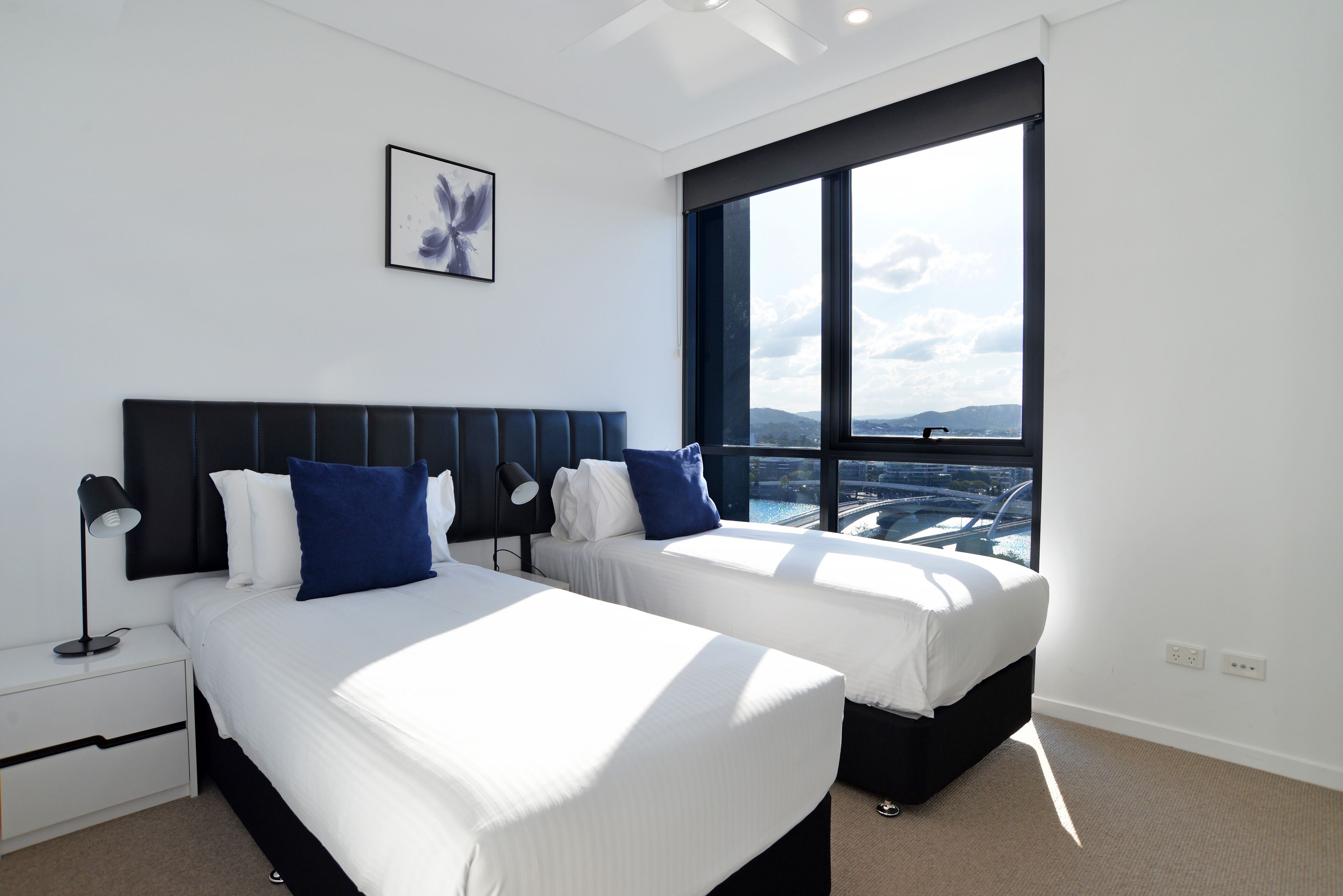 A modern hotel room with two beds, a large window with a city view, a nightstand, and a wall-mounted picture.