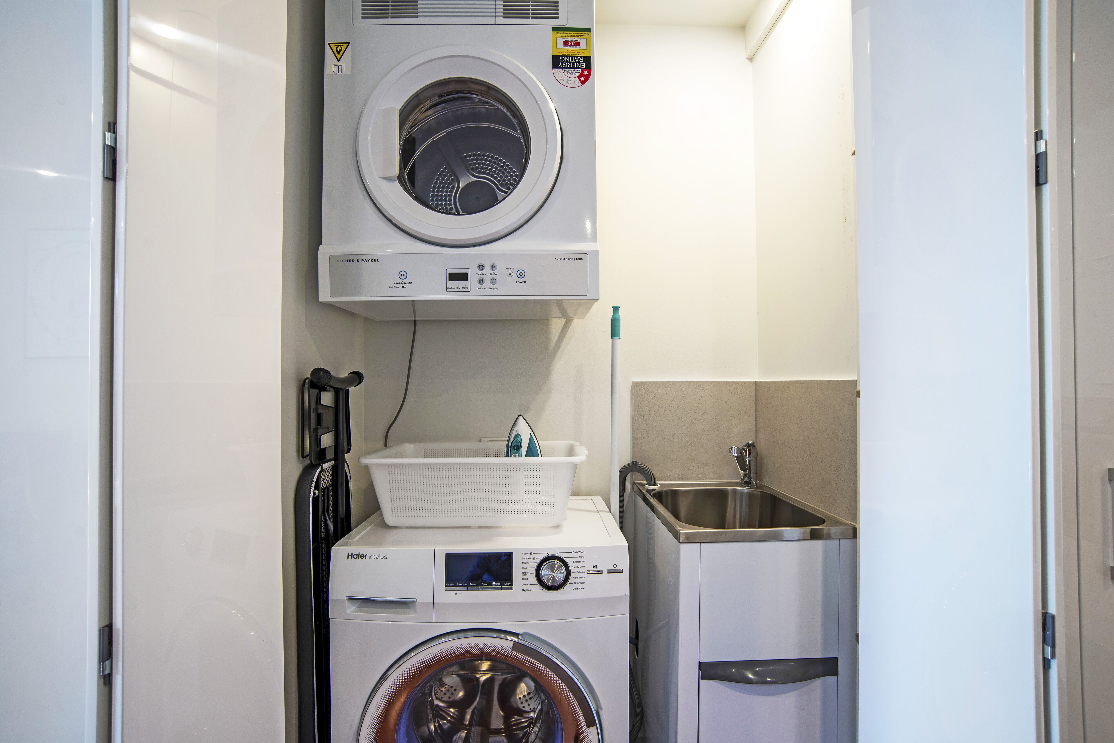 A compact laundry area with a front-loading washer and dryer, a small sink, and a basket on the dryer.