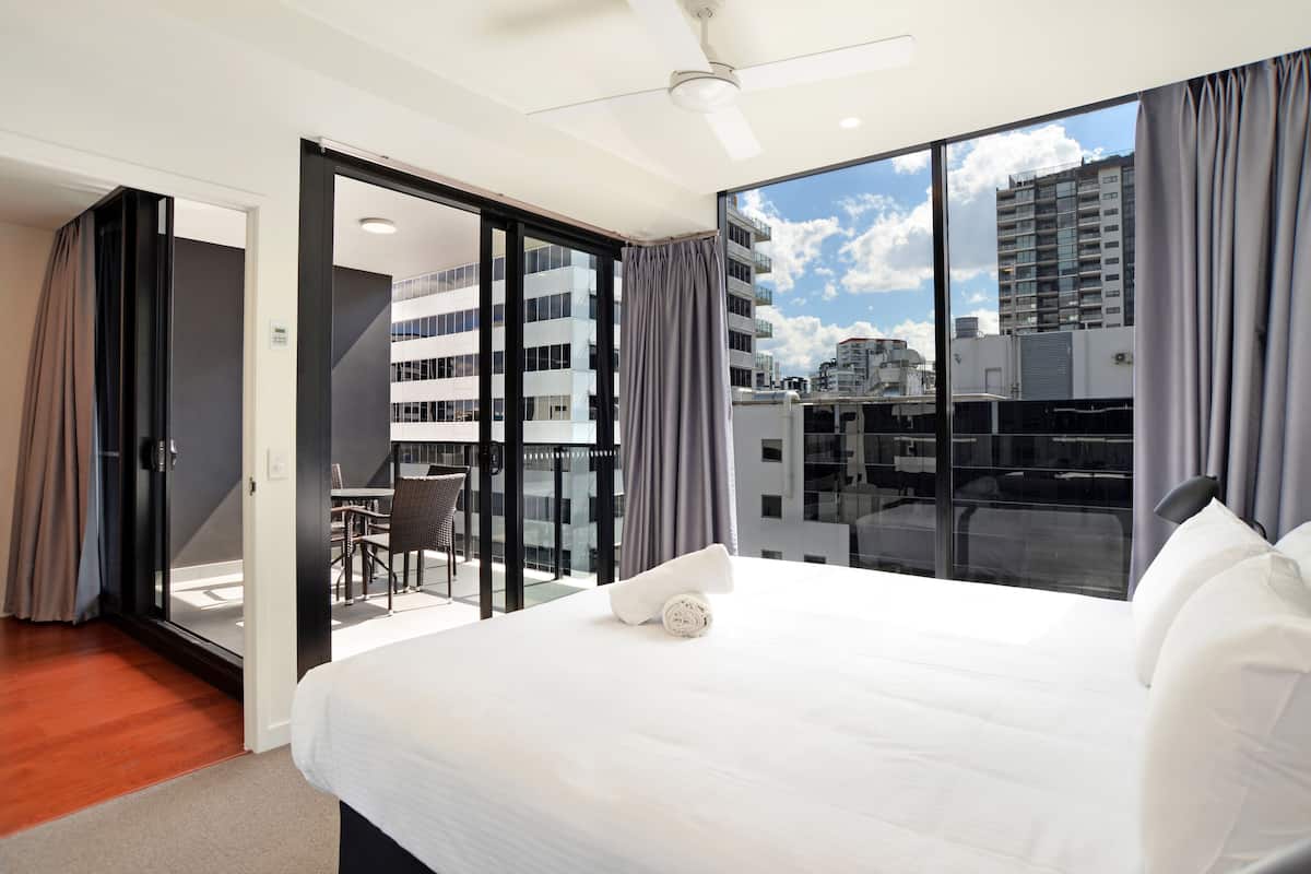 A hotel room with a large bed, a ceiling fan, and a view of the city through large windows.