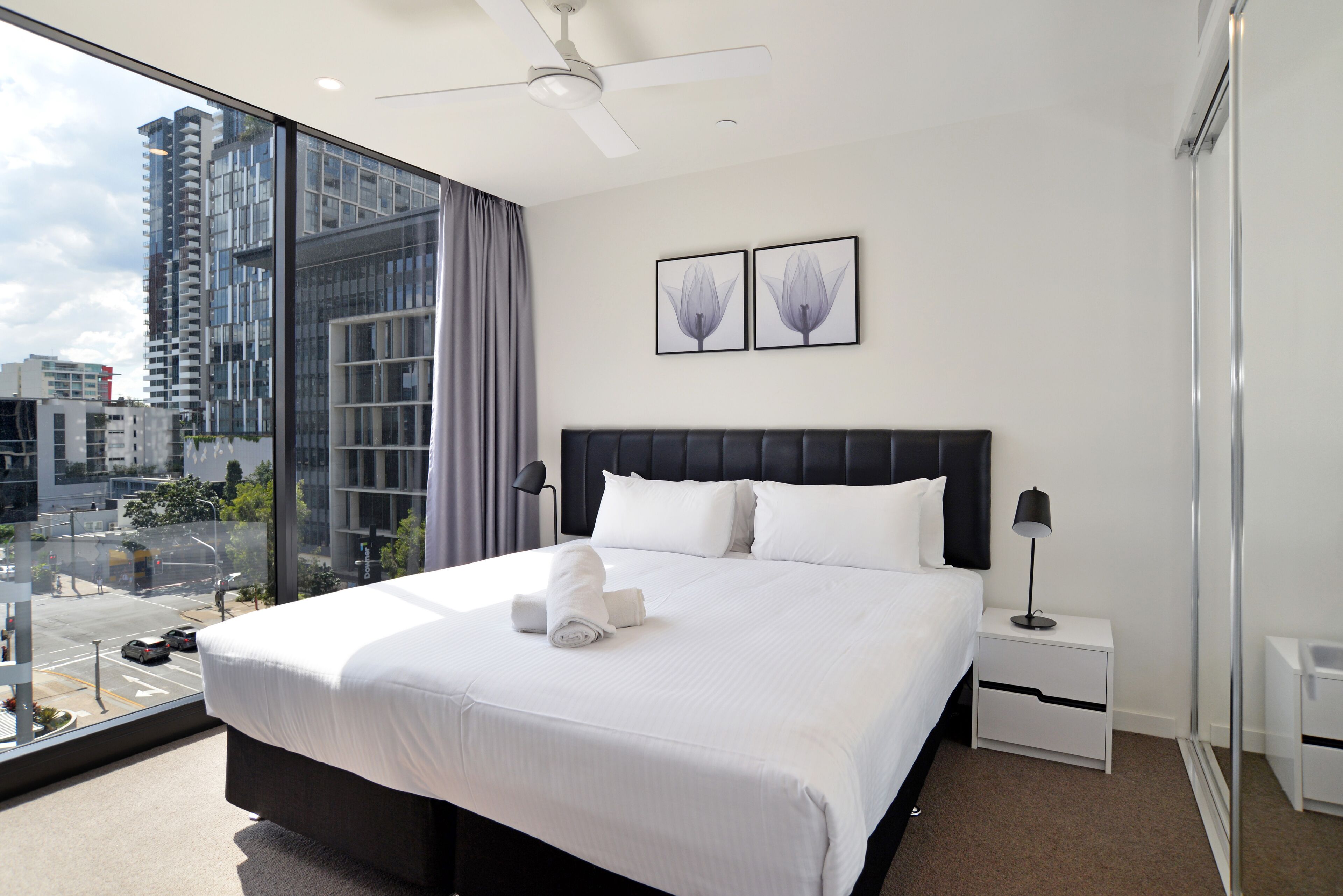 A modern hotel room with a large bed, a ceiling fan, and a city view through floor-to-ceiling windows.