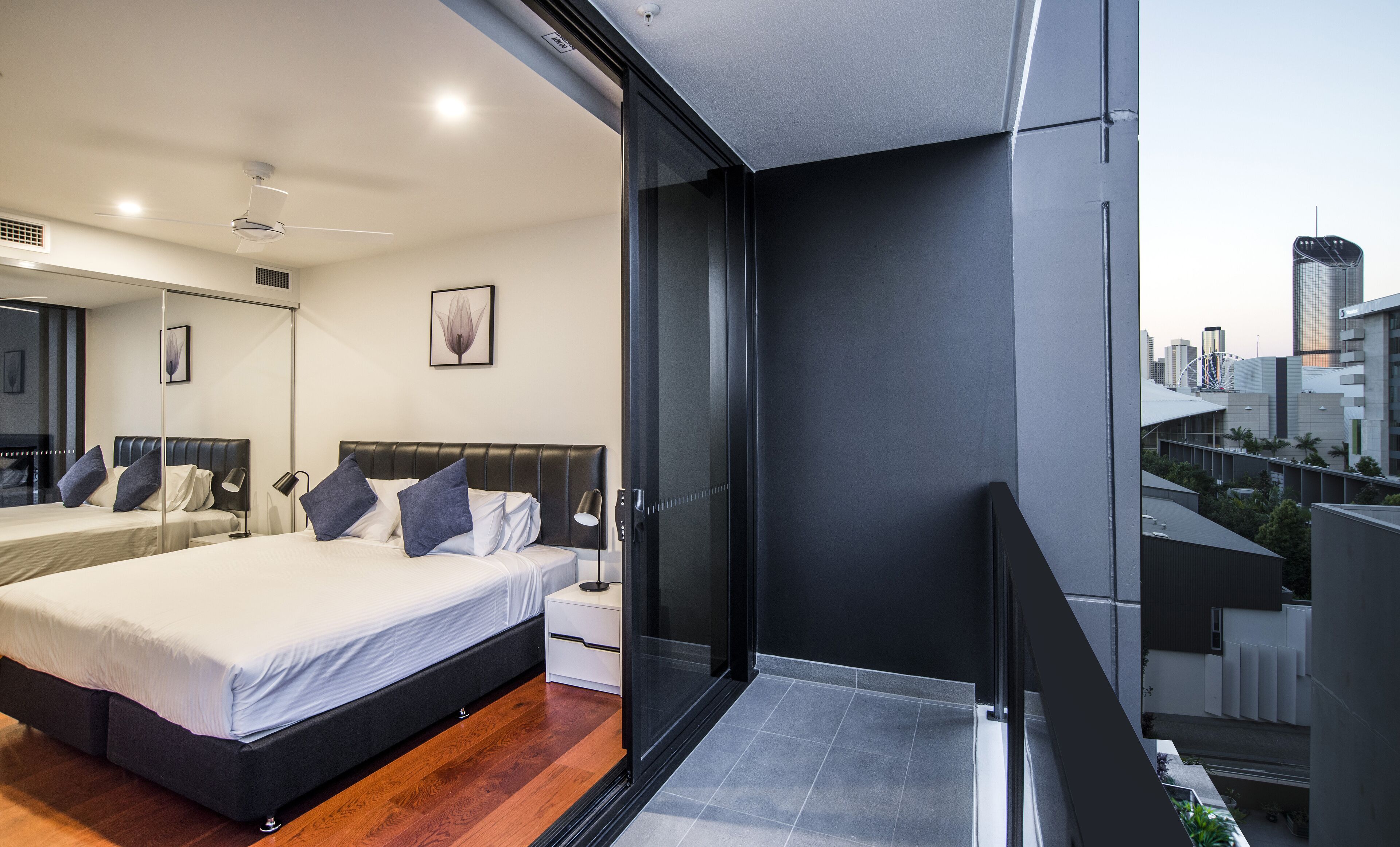 Una habitación de hotel moderna con una cama grande, mesitas de noche, un espejo y un balcón con vistas a la ciudad.