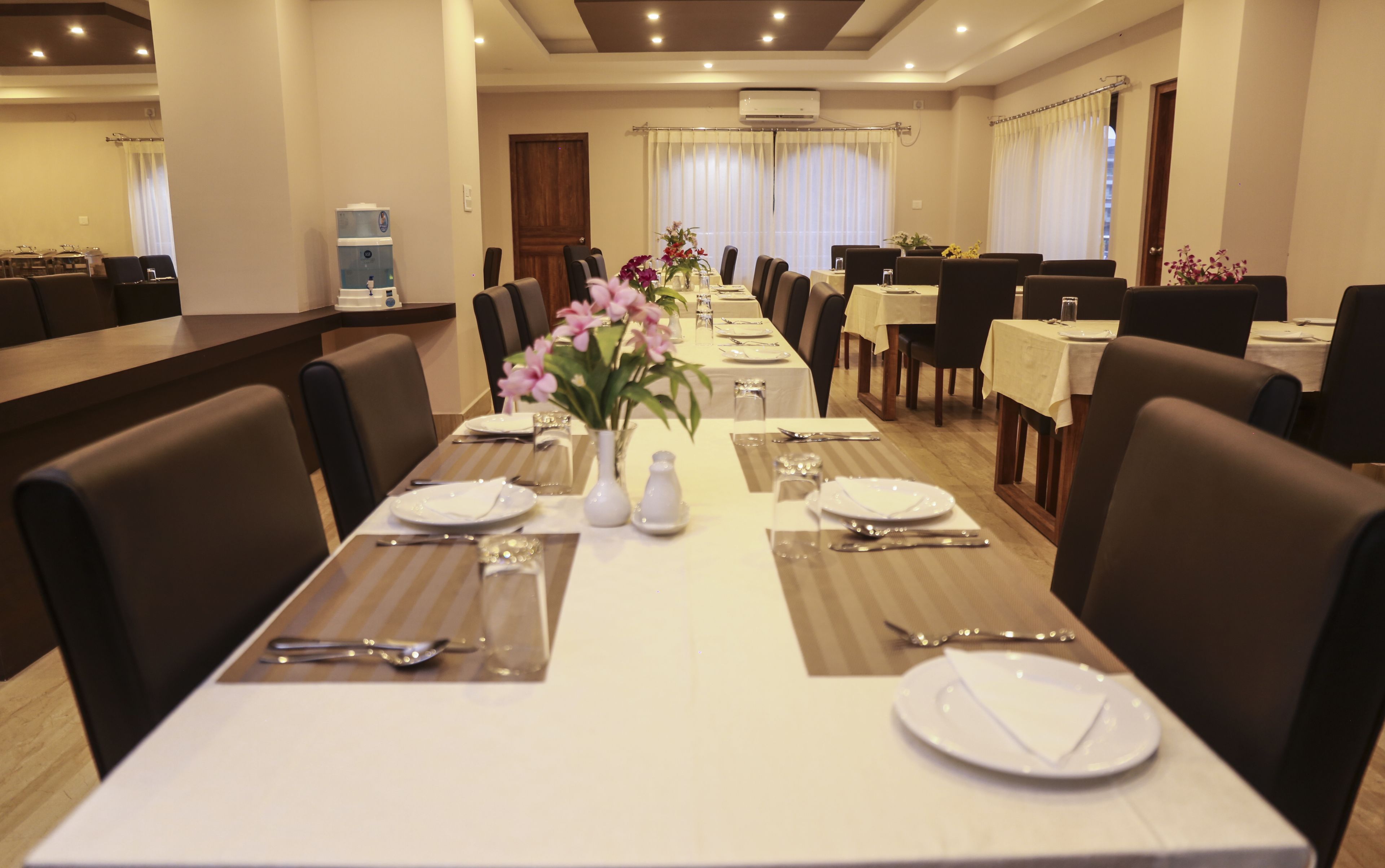 A hotel dining area with multiple tables set for service, a water dispenser, and a floral centerpiece.