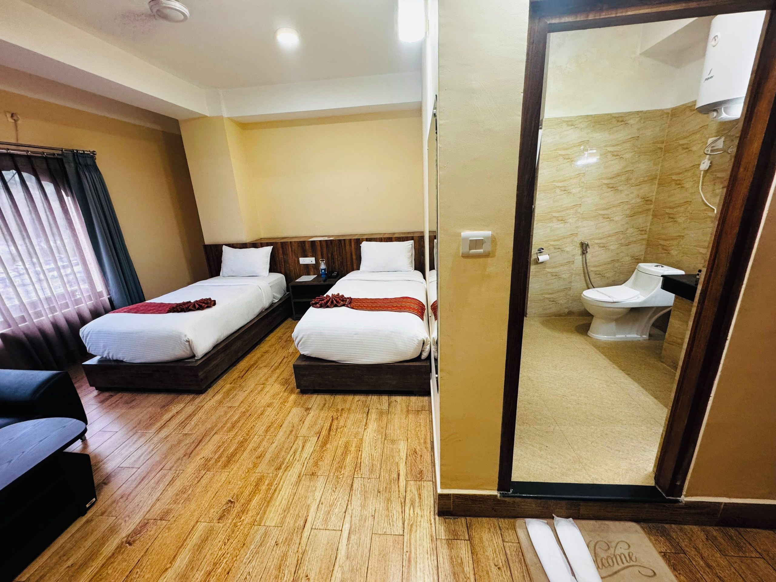 A hotel room with two beds, a wooden floor, a bathroom with a toilet and shower, and a door leading to another room.