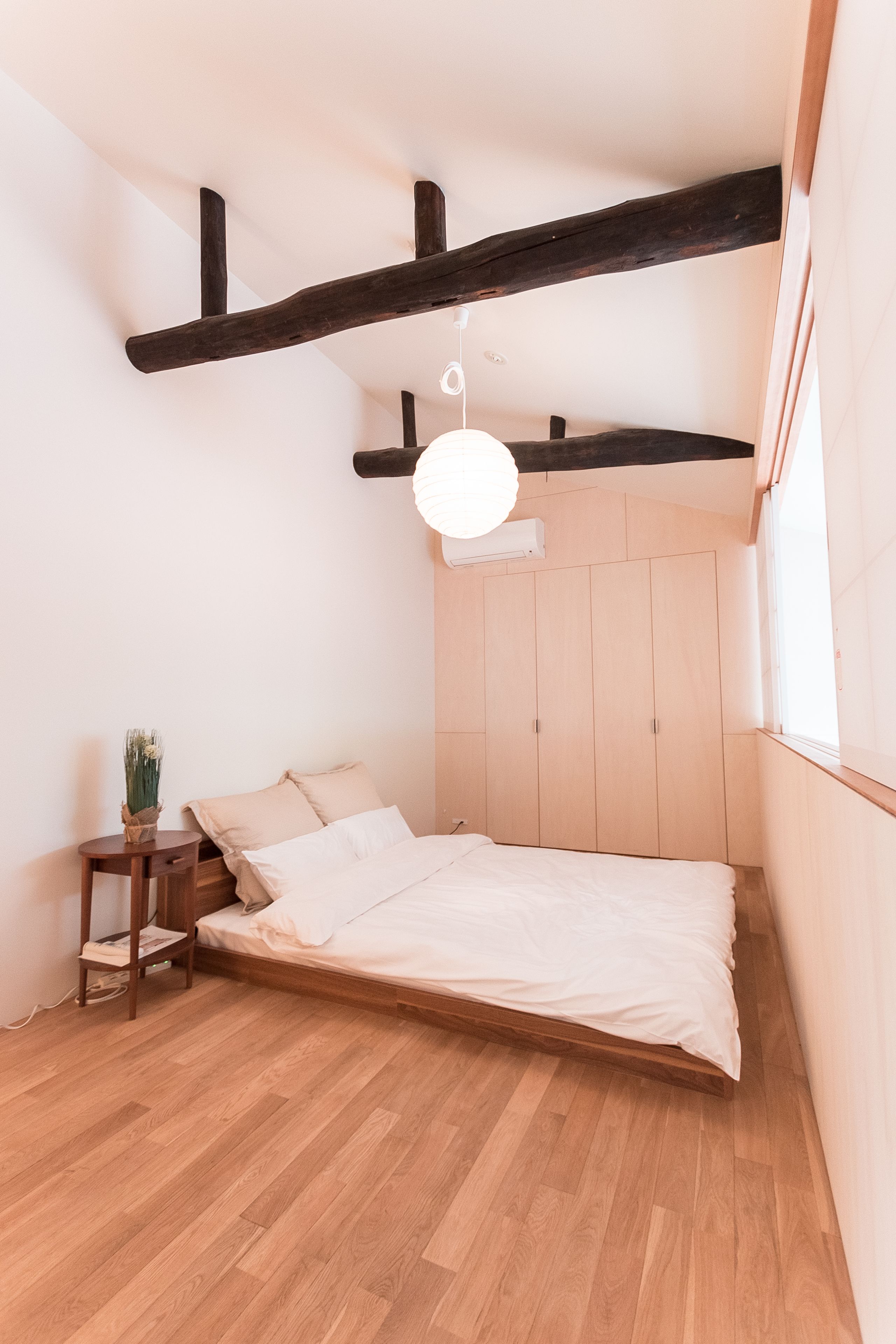 Een slaapkamer met een houten vloer, een bed met wit beddengoed, een klein nachtkastje van hout, een potplant en een hanglamp.
