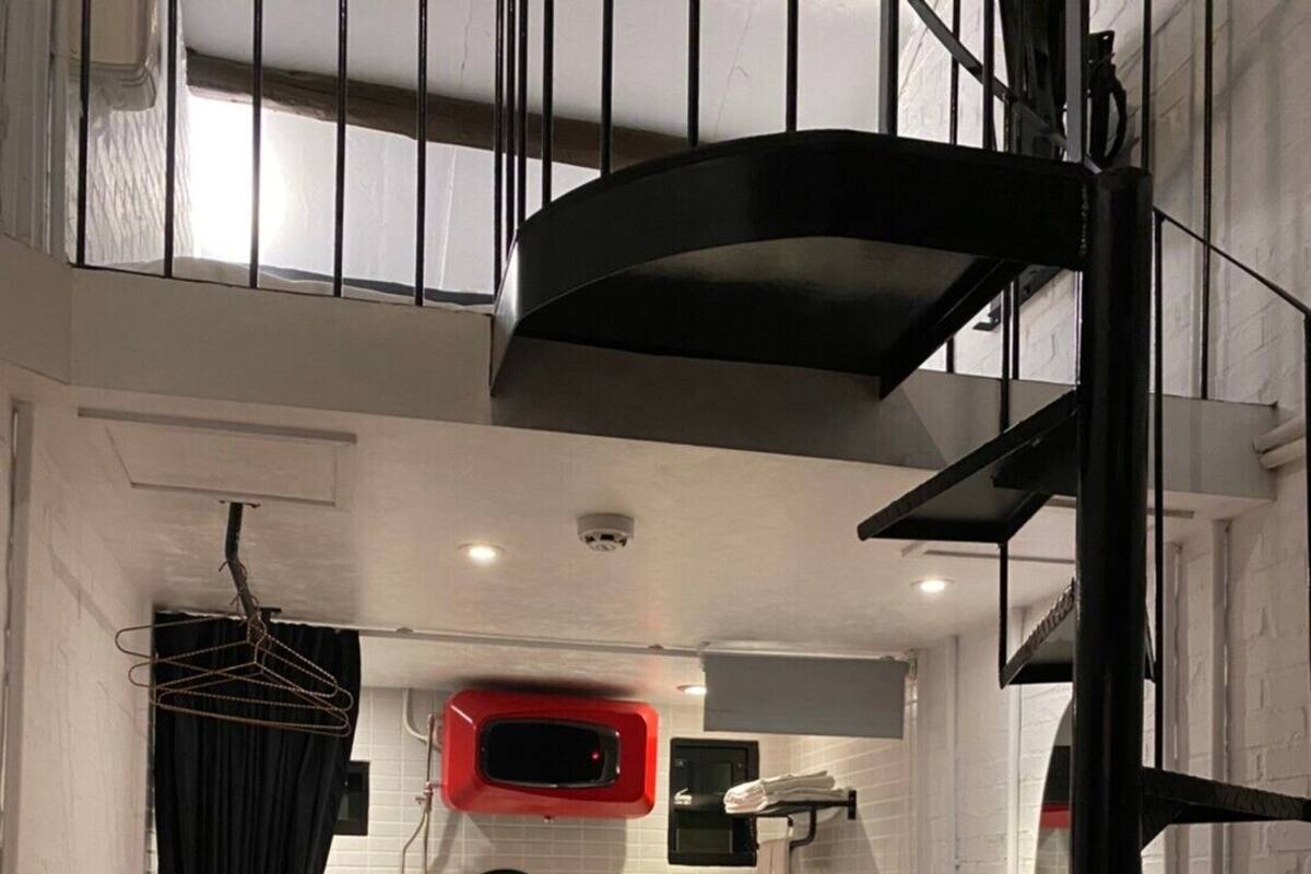 A compact, modern bathroom with a white tiled wall, a red towel rack, a black hanging shower curtain, a white toilet, and a wooden chair.