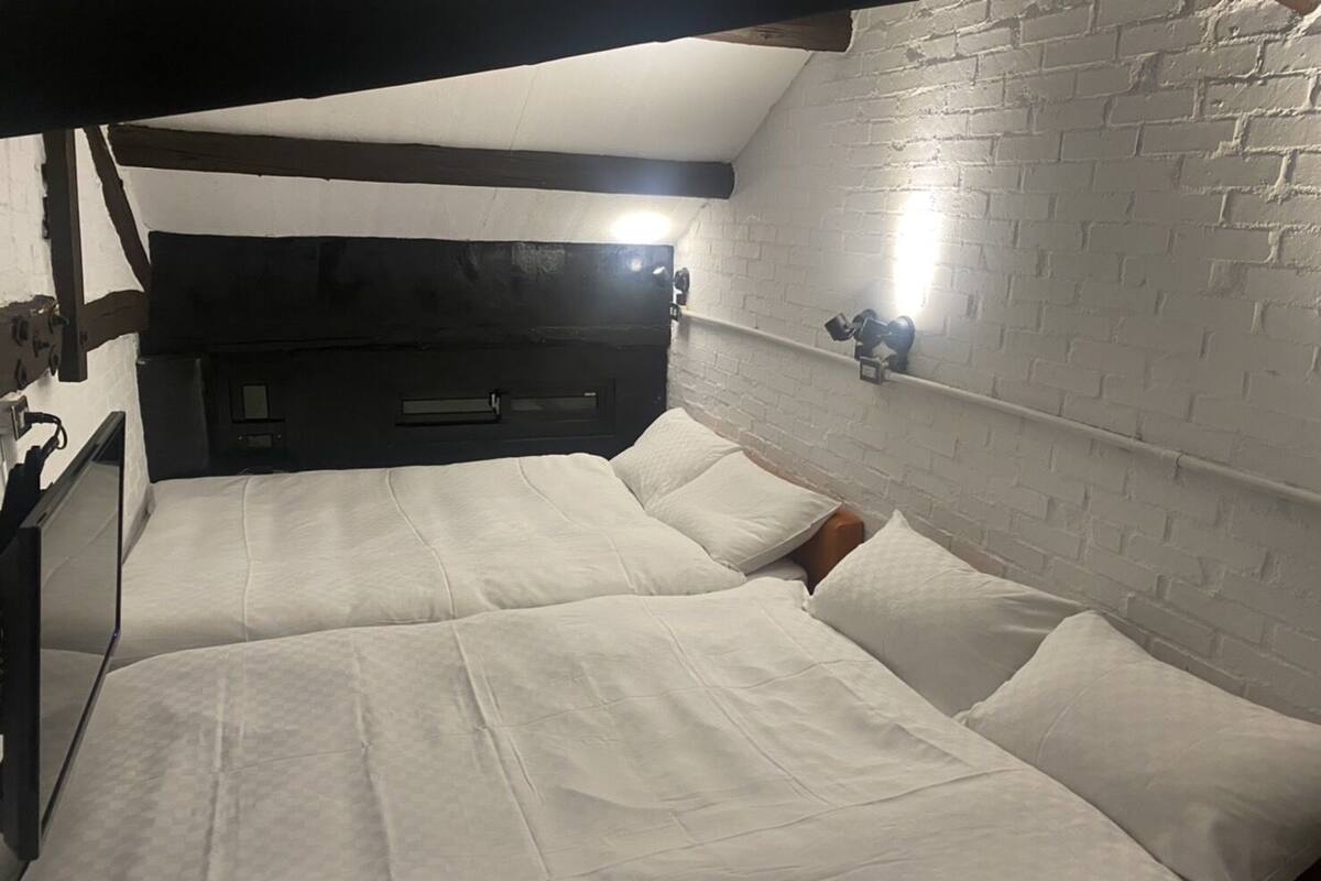 A room with two beds, white bedding, a brick wall, and a shelf with a lamp.