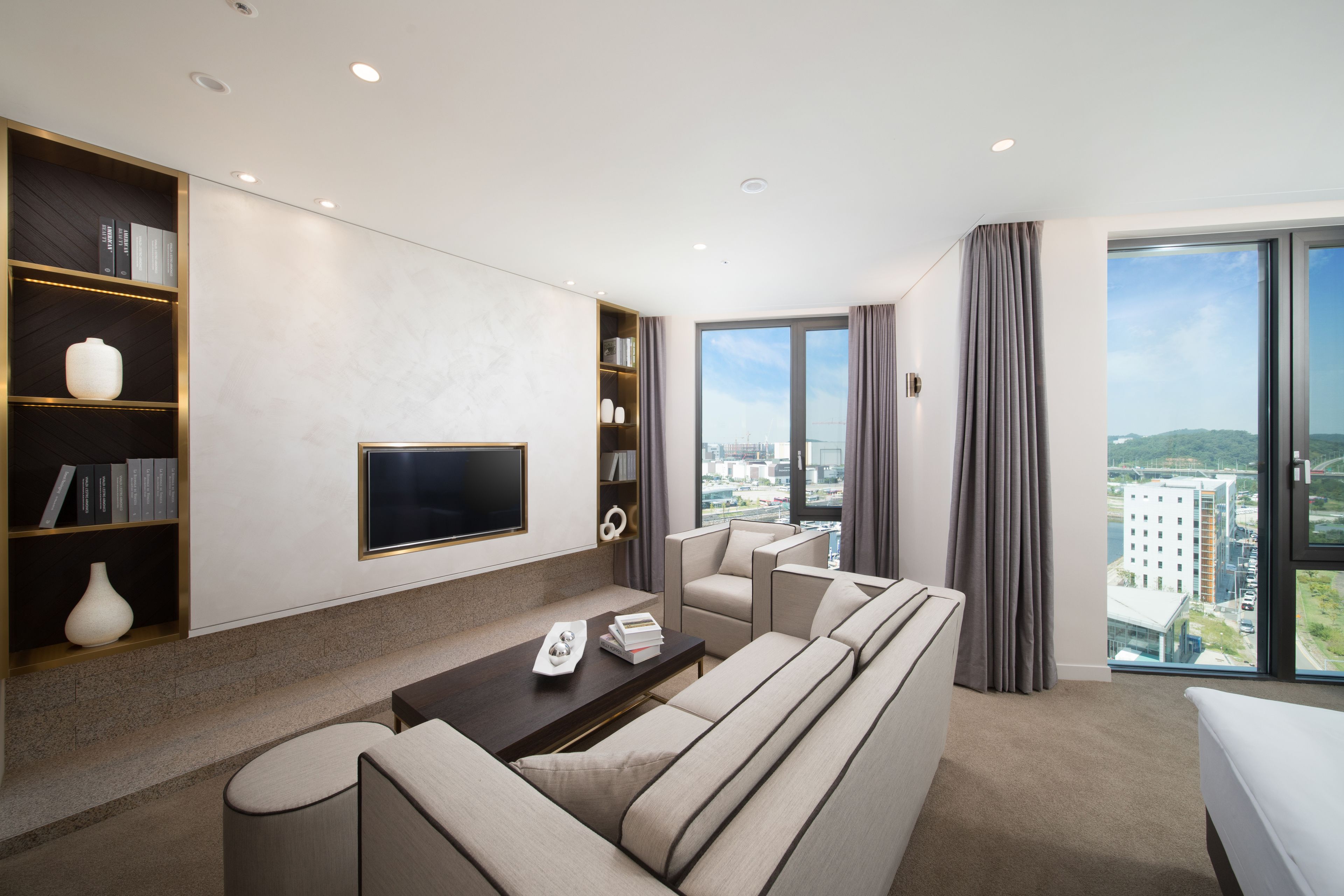 A modern living room with a sofa, armchairs, a coffee table, and a flat-screen TV mounted on the wall. There are bookshelves with books and decorative items, and a large window offering a city view.