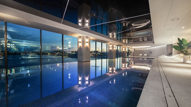 Une piscine intérieure moderne aux parois en verre transparent, offrant une vue sur le paysage urbain au crépuscule.
