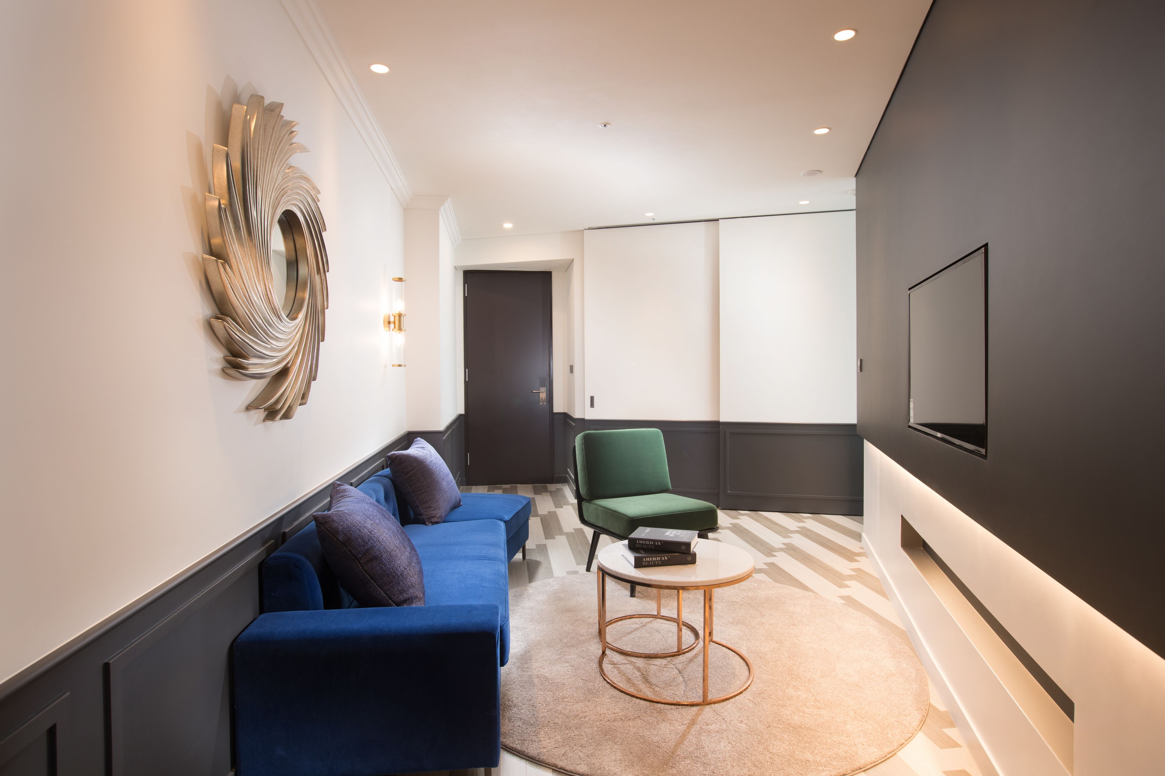 Een moderne woonkamer met een blauwe bank, een groene fauteuil, een ronde salontafel en een grote flatscreen televisie aan de muur.
