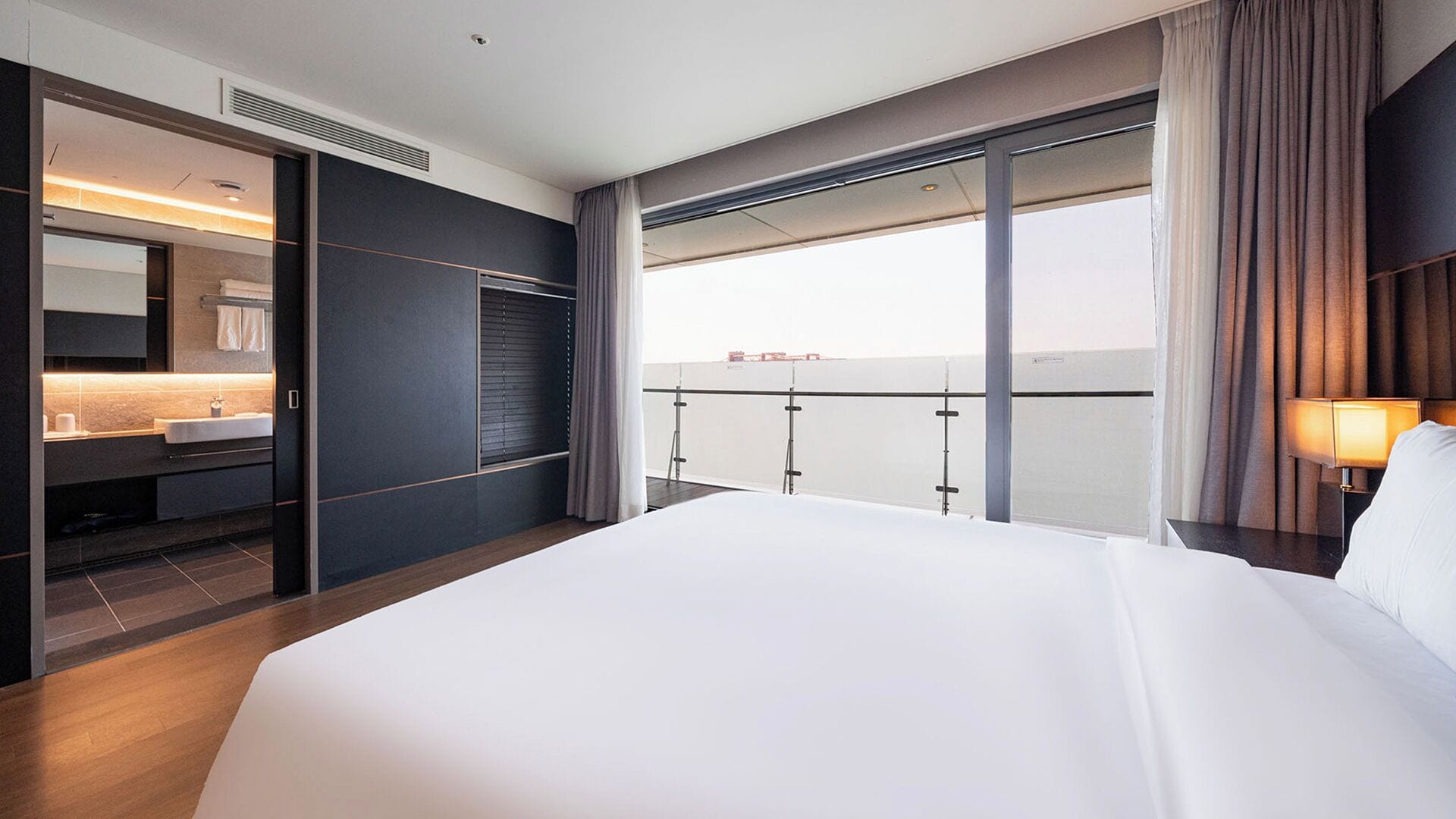 Una habitación de hotel moderna con una cama grande, un baño con tocador y un balcón con vistas a la ciudad.