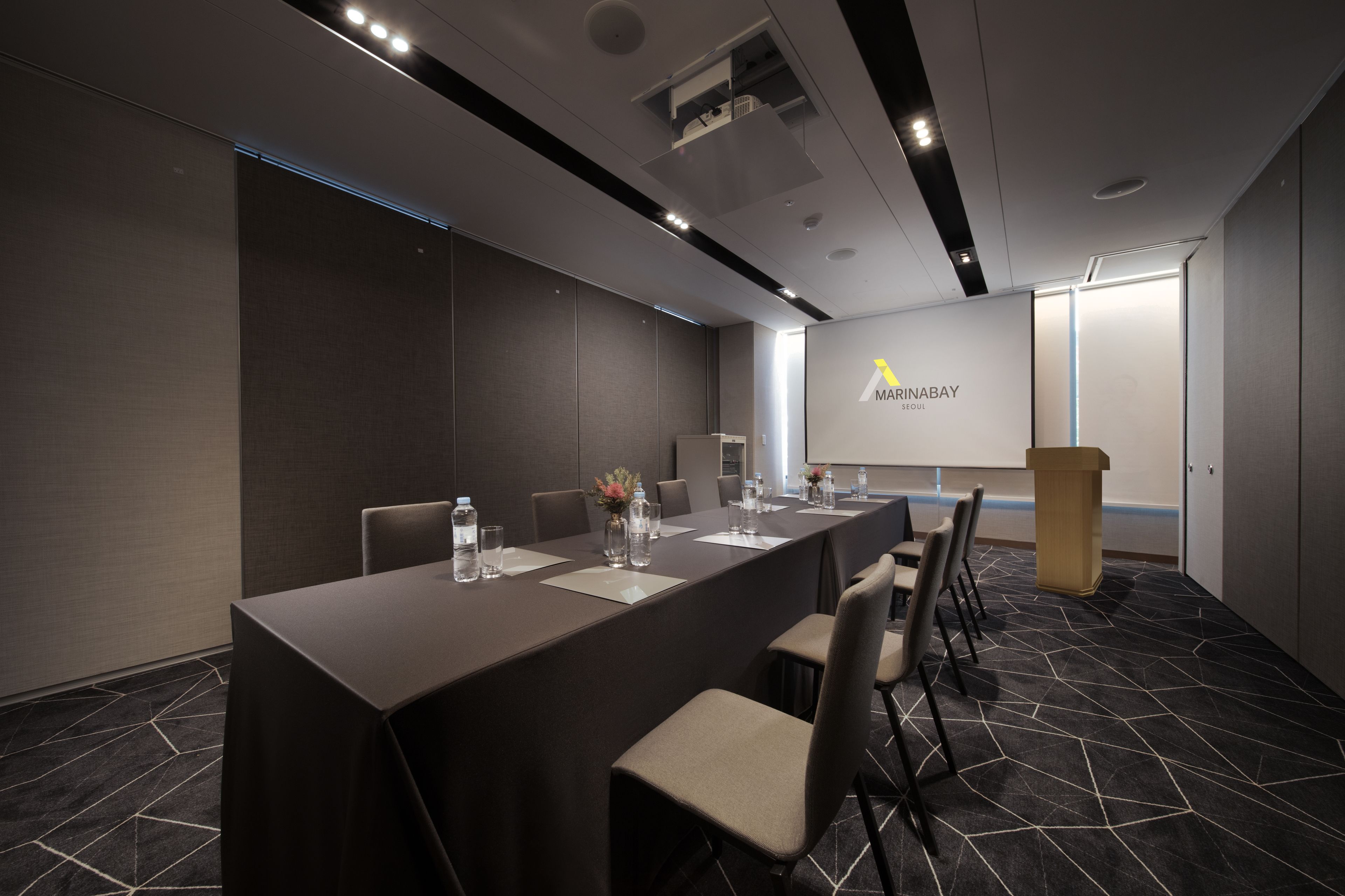 Un salón de conferencias con una larga mesa, sillas, un atril y una pantalla para presentaciones.