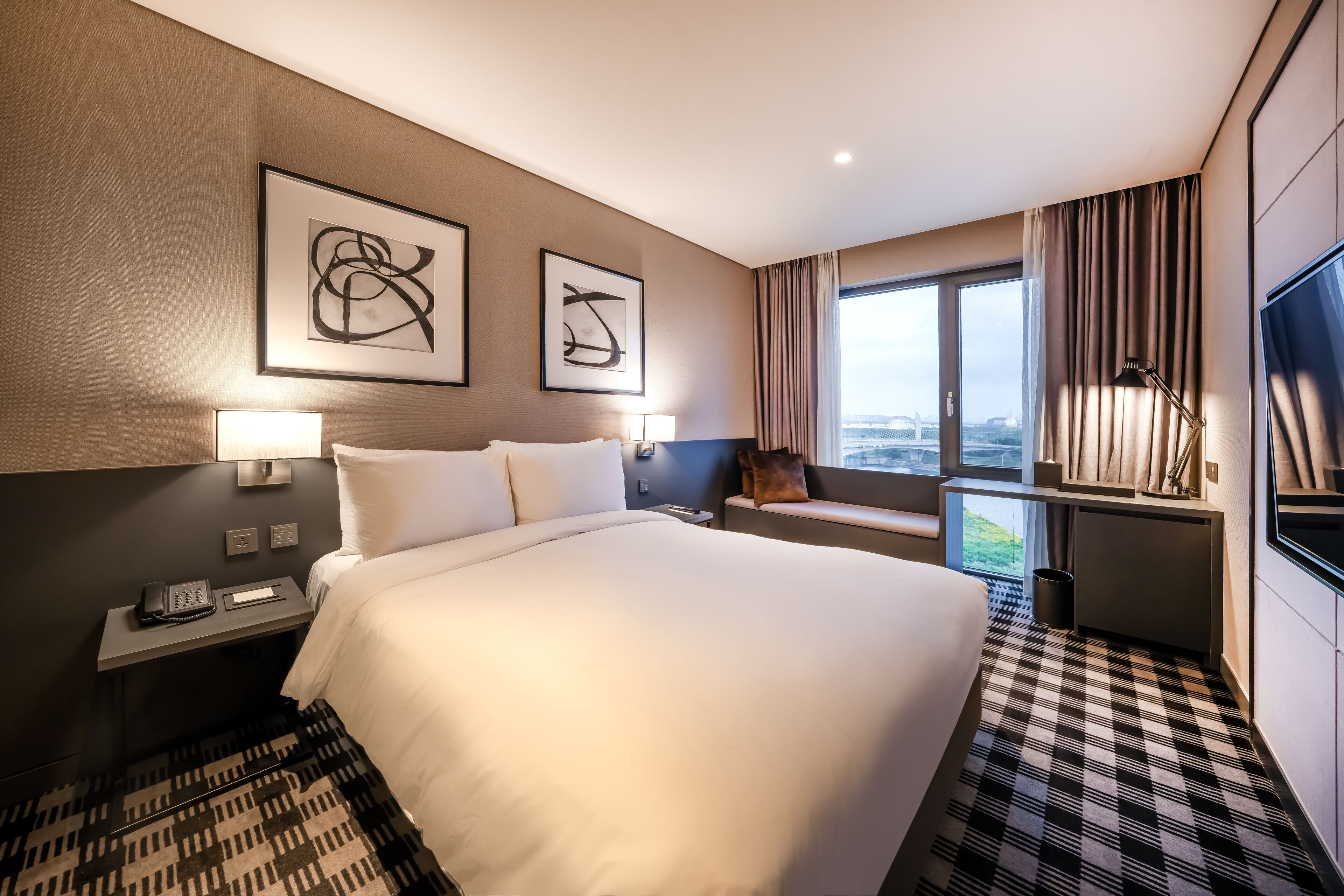 A hotel room with a large bed, two bedside tables with lamps, a desk with a chair, a TV, and a window with curtains.