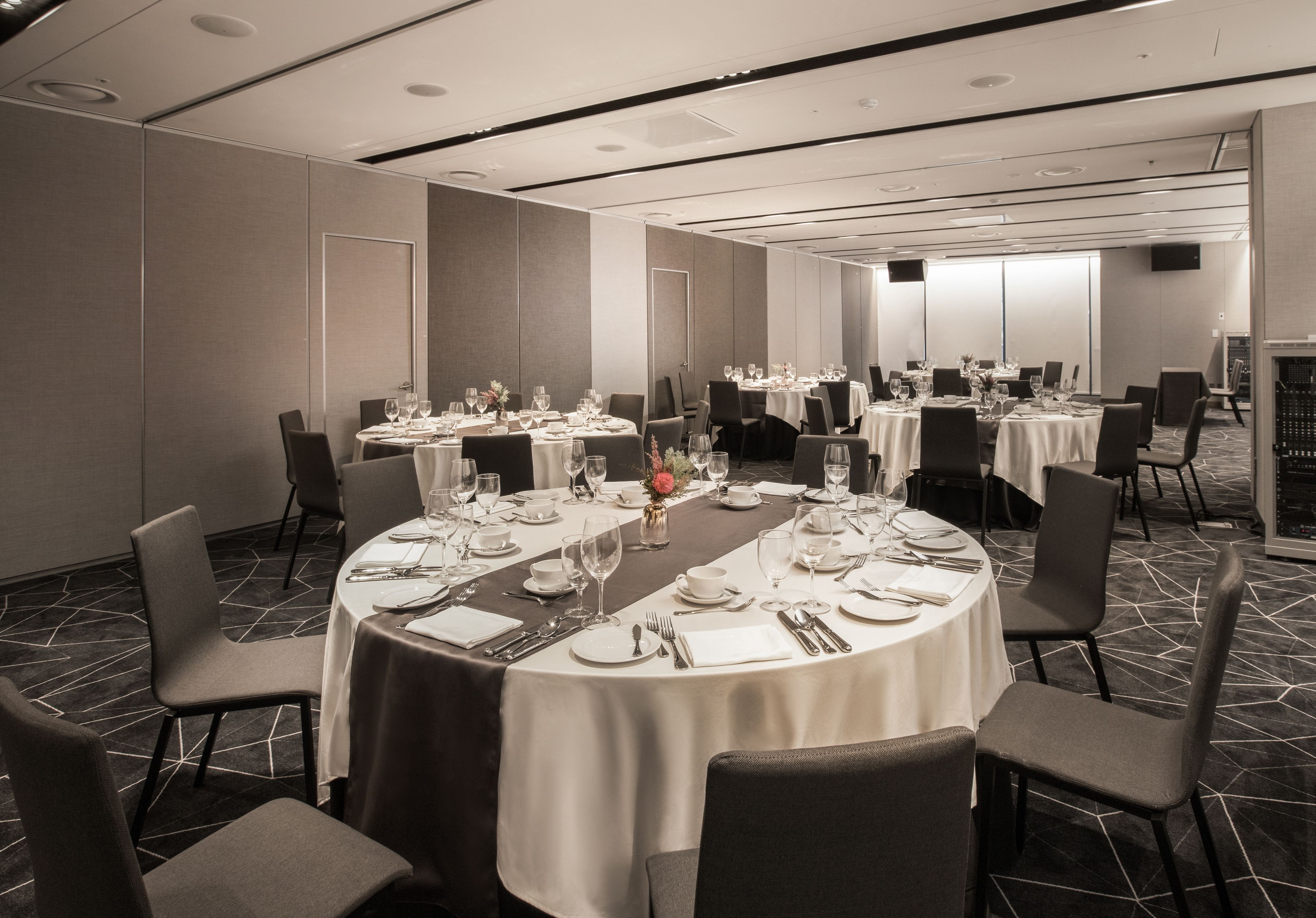 Sala de conferências com mesas redondas preparadas para refeições, equipada com cadeiras, pratos, talheres e copos.