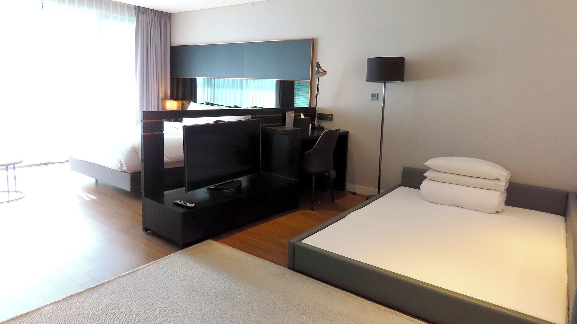 A modern hotel room with a bed, a desk, a chair, a TV, and a lamp.