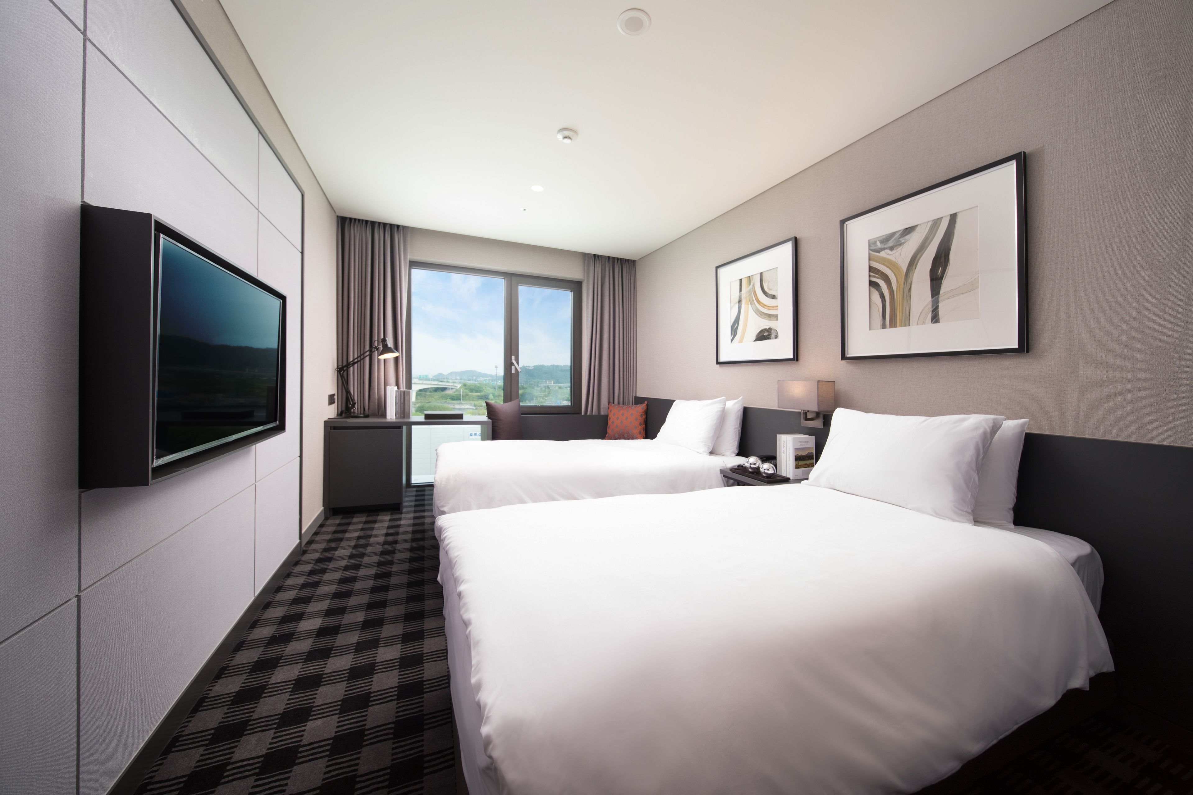 A hotel room with two beds, a flat-screen TV, artwork on the walls, and a large window with a view.