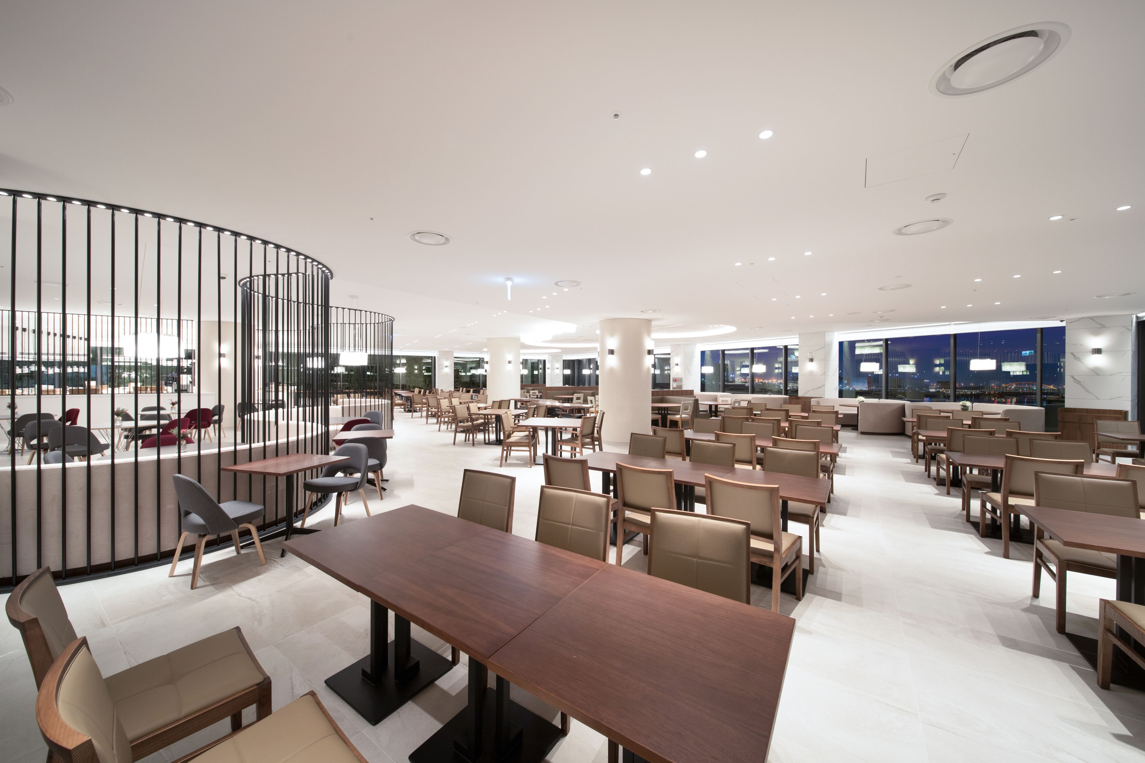 A spacious indoor dining area with multiple tables and chairs, a curved black railing, and large windows offering a view of the cityscape.