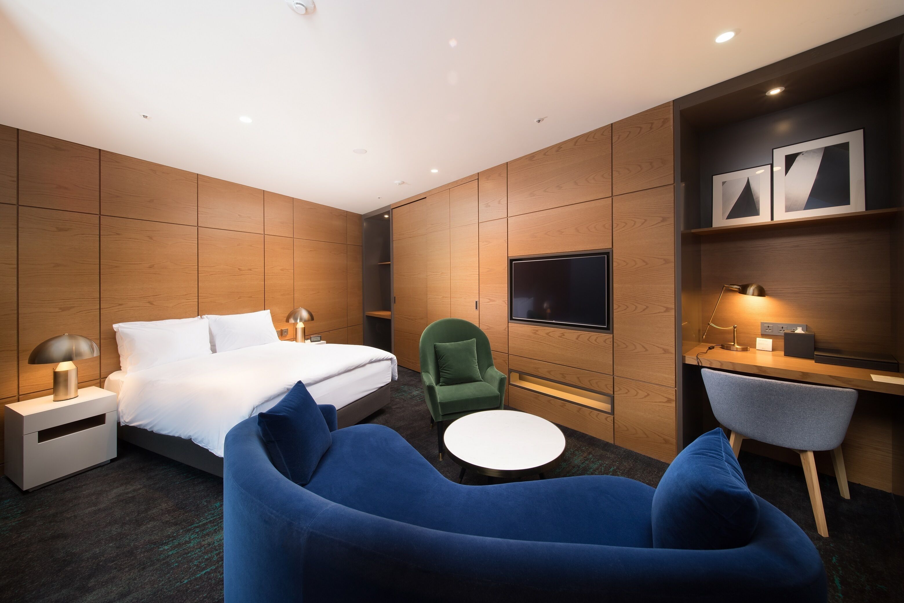 A modern hotel room with a bed, a desk, a TV, and seating area.