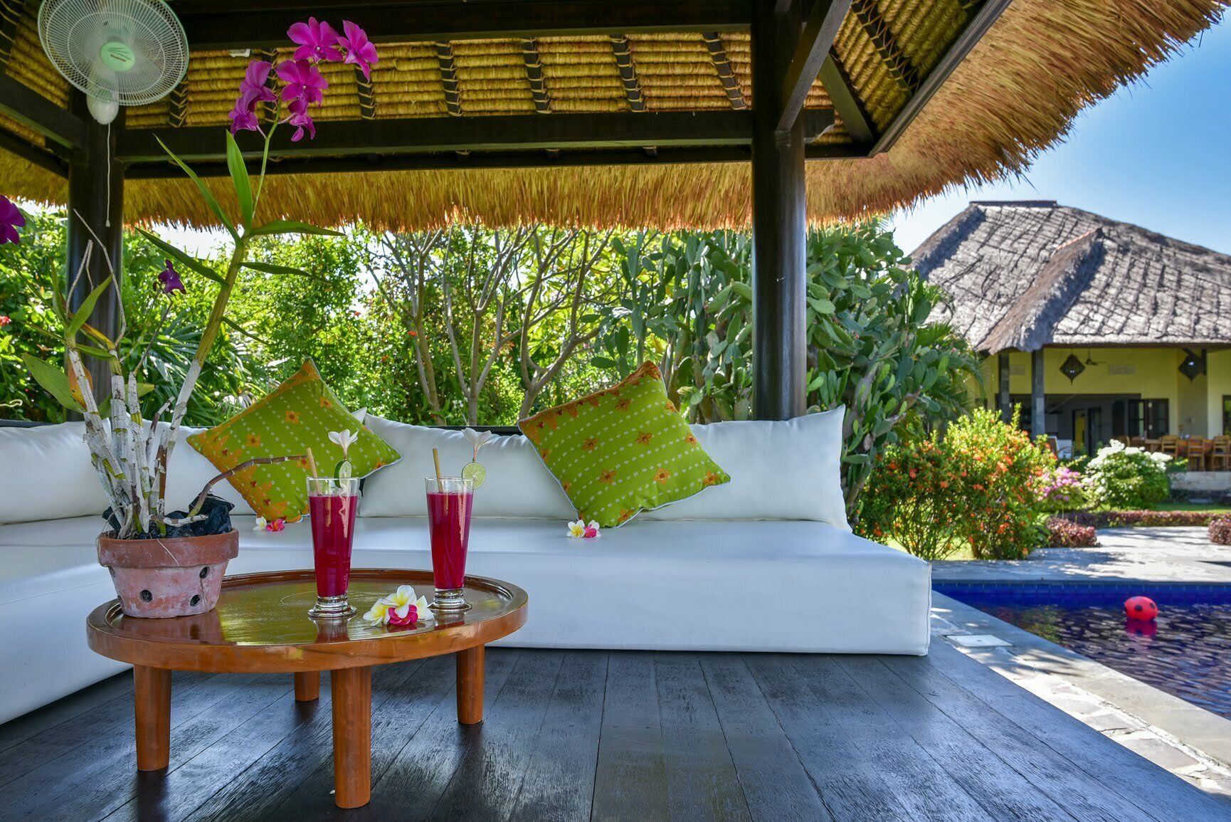 A tropical outdoor seating area with a thatched roof, a pool, and lush greenery.