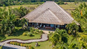 A thatched-roof building with a patio and outdoor seating area surrounded by lush greenery and palm trees. - Direct beachfront 4BR/BR villa in tropical garden. Villa chef, villa pool, WiFi (Umeanyar)