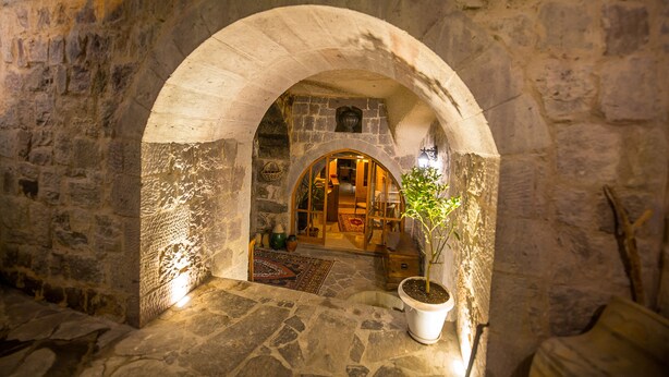 An arched stone doorway leading into a room with a wooden door, a potted plant, and a rug on the floor. - Maya Cave Hotel (Nevsehir)