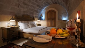 A cozy room with a stone ceiling, a neatly made bed, a dining table with plates of fruit and a wine glass, and a candle on the table. - Maya Cave Hotel (Nevsehir)