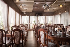 Un espace repas avec des tables et des chaises en bois, de grandes fenêtres avec des rideaux et des lampes suspendues.
