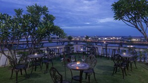 An outdoor dining area with round tables and chairs, overlooking a river and cityscape at dusk. - Le Castle River Hotel & Apartment (Phnom Penh)