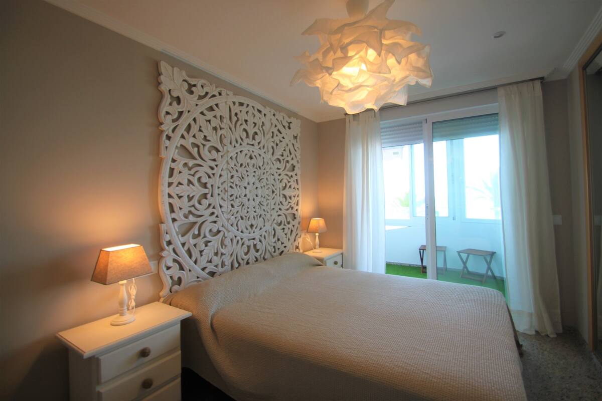 A bedroom with a large bed, two bedside tables with lamps, a decorative wall panel, and a view of a balcony with a table and chairs.