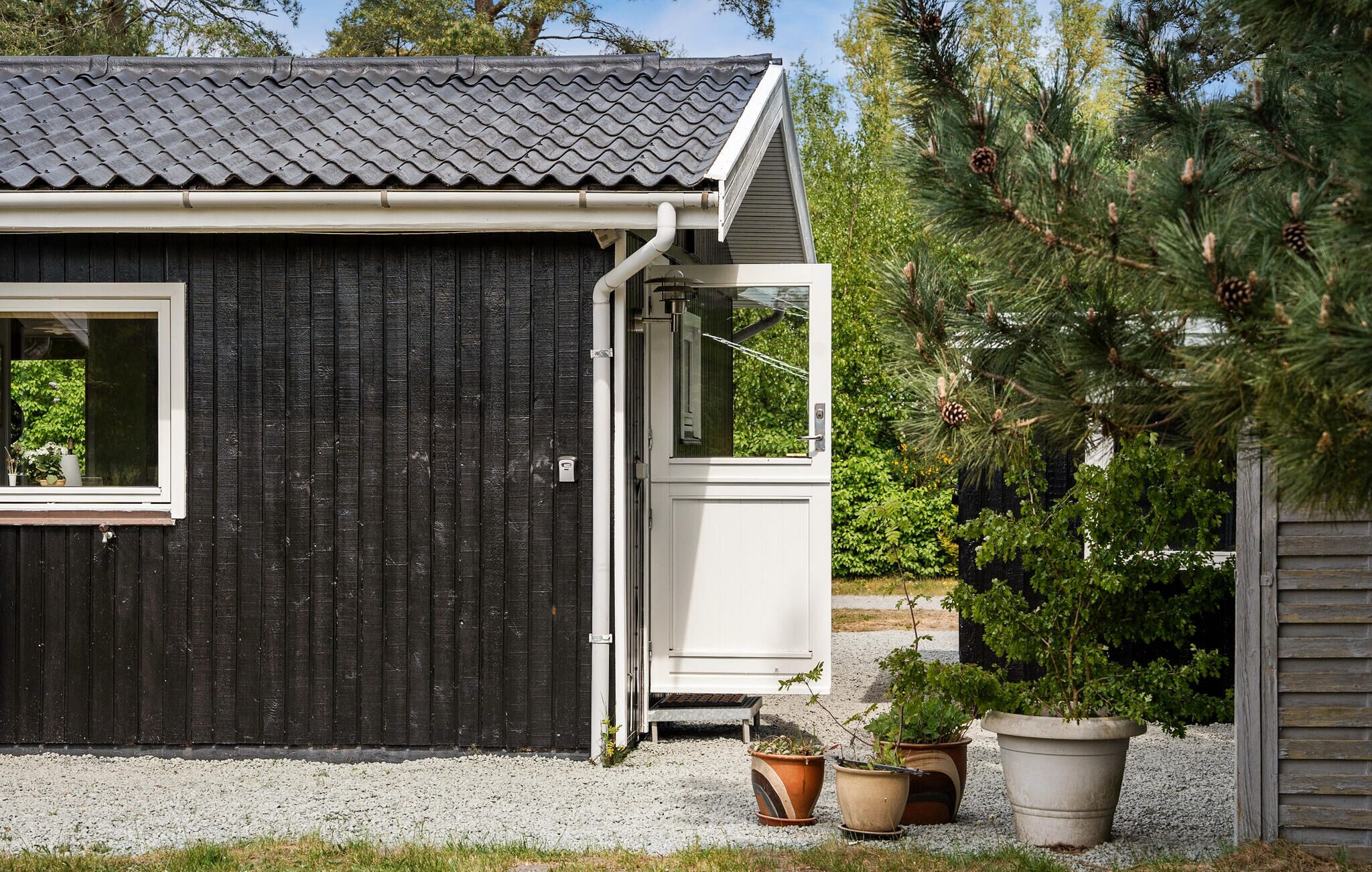 Un petit bâtiment en bois sombre, avec un toit en tuiles, une porte blanche et une fenêtre. Des plantes en pot sont disposées devant le bâtiment.