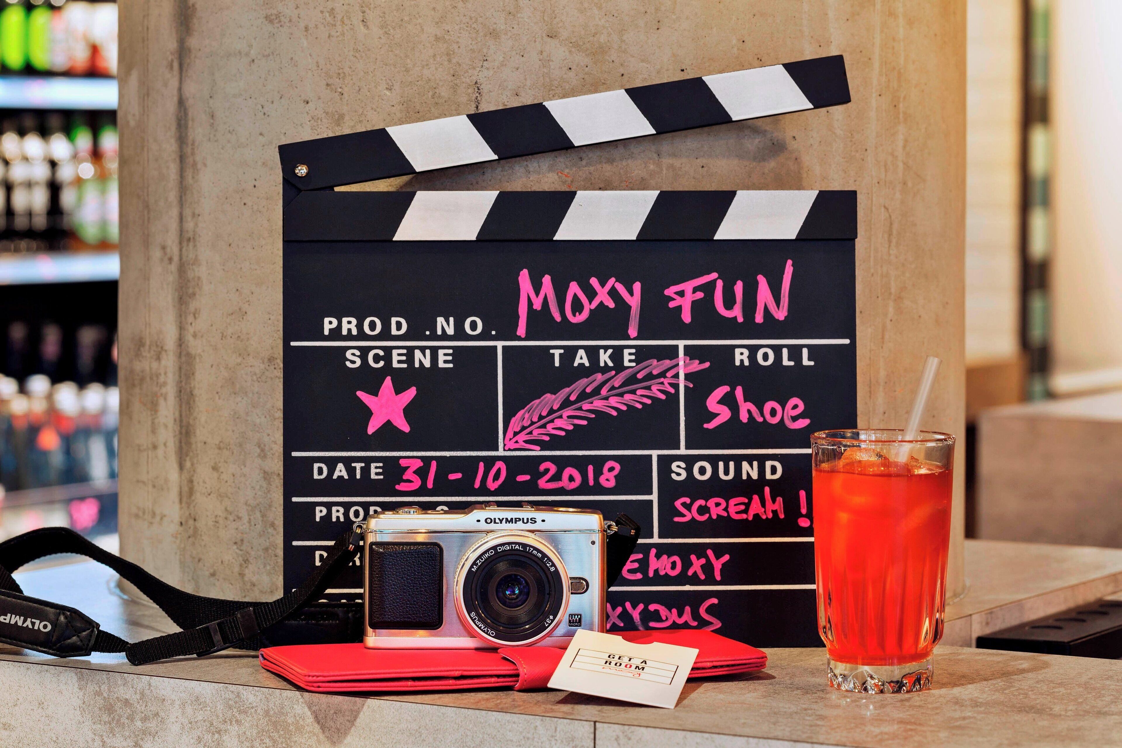 A clapperboard with handwritten notes, a vintage Olympus camera, and a glass of red beverage on a table.