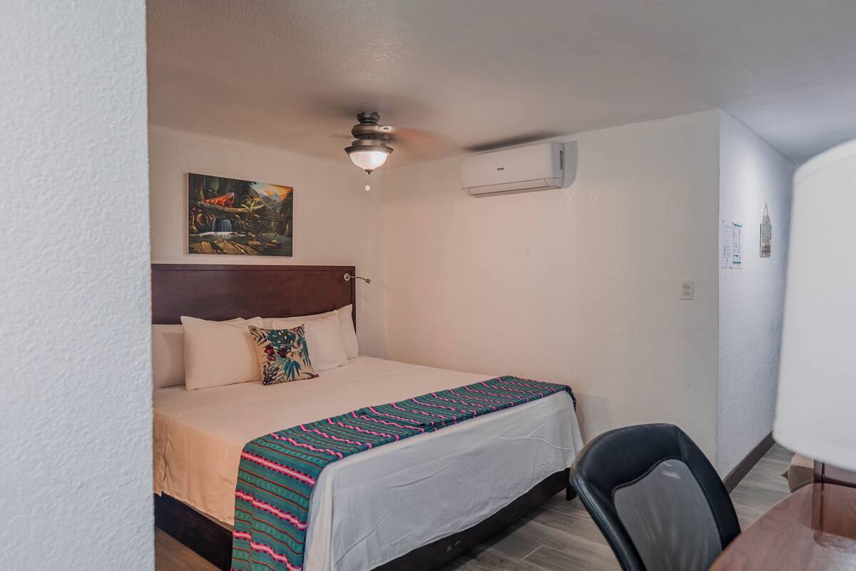 A hotel room with a bed, a desk, a chair, a ceiling fan, an air conditioning unit, and a painting on the wall.