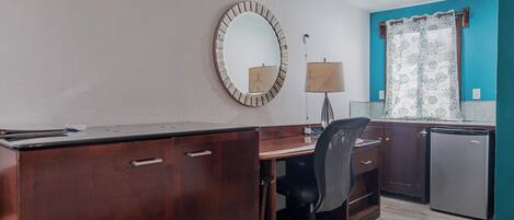A compact office space with a wooden desk, a chair, a small refrigerator, and a mirror on the wall.