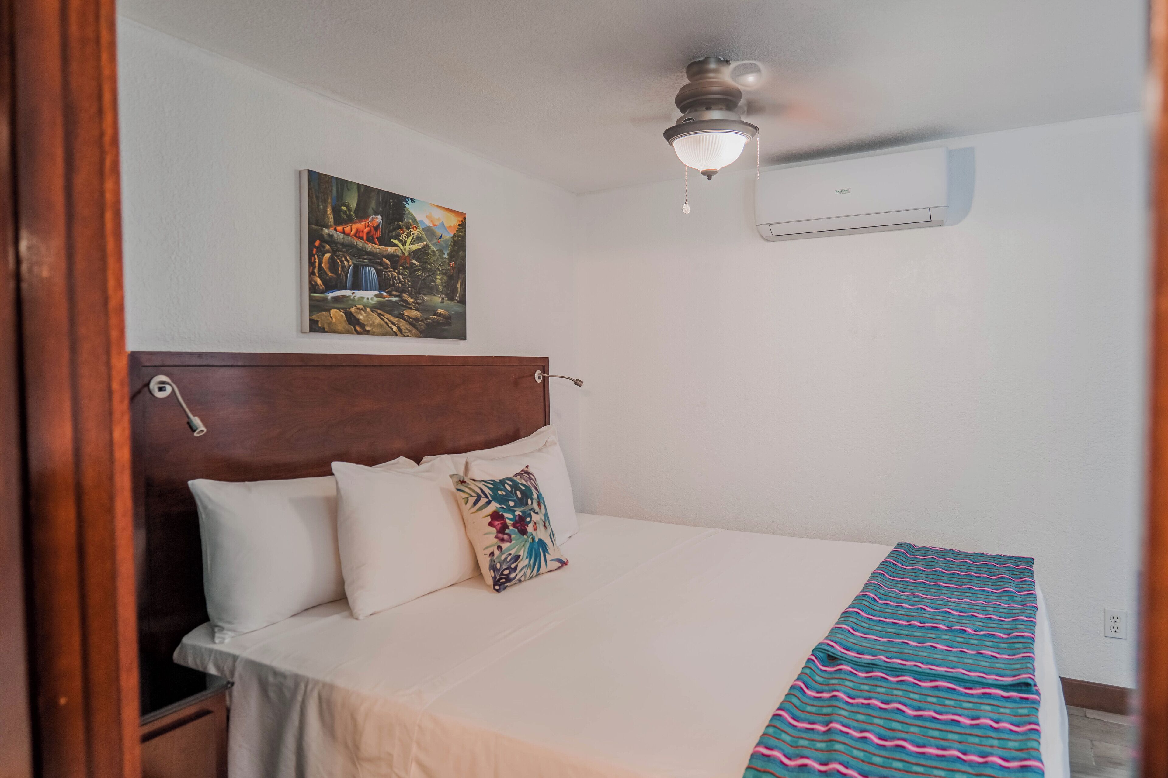 A bedroom with a wooden headboard, a bed with white bedding, a colorful pillow, a ceiling fan, and a wall-mounted air conditioner.