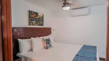 A bedroom with a wooden headboard, a bed with white bedding, a colorful pillow, a ceiling fan, and a wall-mounted air conditioner.