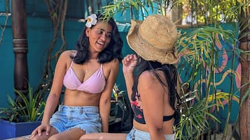 Dos personas sentadas junto a una piscina, con follaje y una pared azul al fondo.