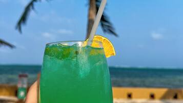 A hand holding a green drink with a straw and a slice of orange on a beach with palm trees and the ocean in the background.