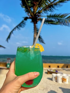 A hand holding a green drink with a straw and a slice of orange on a beach with palm trees and the ocean in the background. - Drift Inn San Pedro (San Pedro)