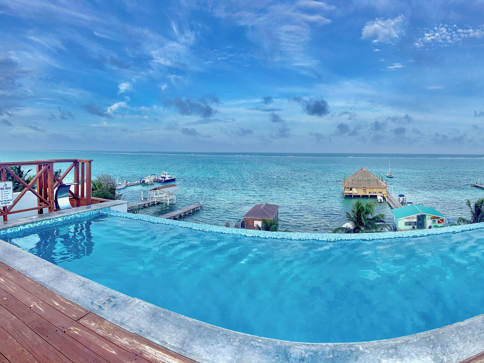 Piscina azul cristalina con terraza de madera, con vistas al océano, bungalows sobre el agua y embarcaciones.