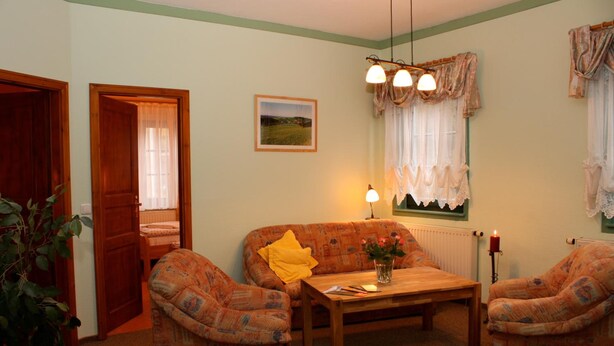 A cozy living room with a sofa, two armchairs, a coffee table, and a lamp. There is a picture on the wall and a window with curtains.