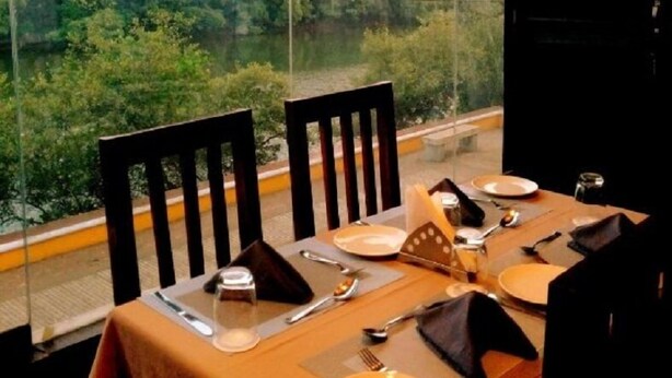 Une table à manger dressée pour deux personnes, avec vue sur une zone boisée grâce à de grandes fenêtres.