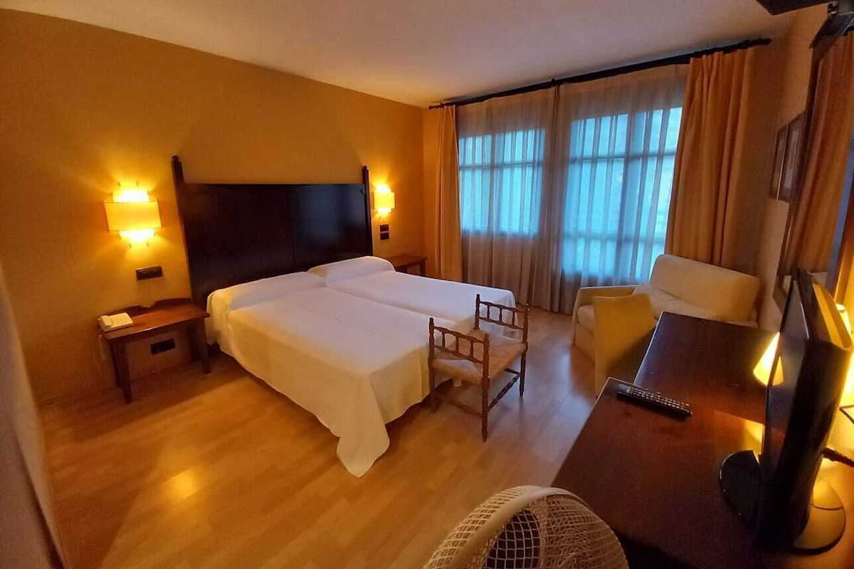 A hotel room with two beds, a desk, a chair, a fan, and a TV.