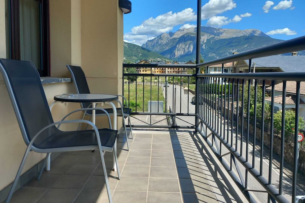 A balcony with two chairs and a small table, overlooking a street and mountains.