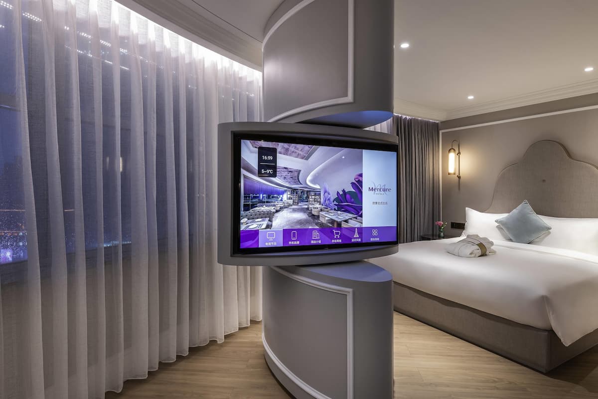 A hotel room with a large flat-screen TV mounted on a curved wall, a bed with white linens, a nightstand, and a view of the city through sheer curtains.