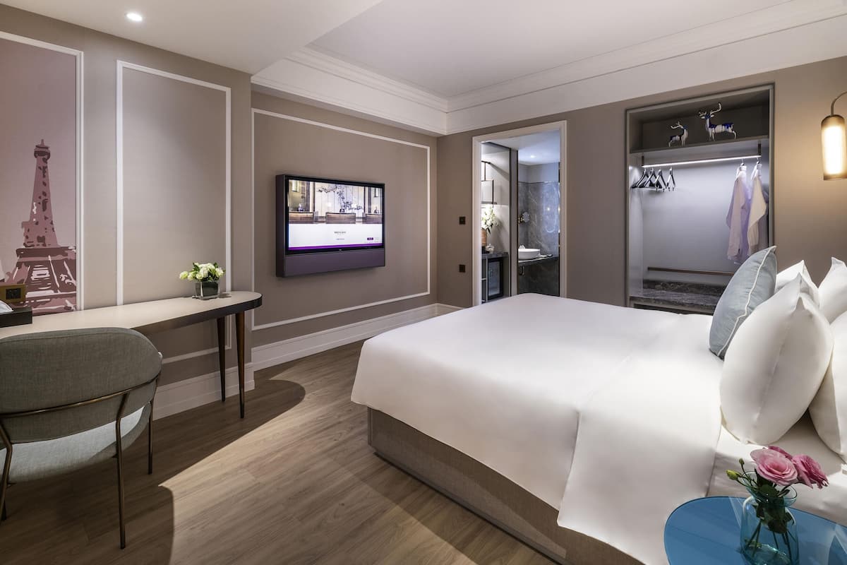 A modern hotel room with a large bed, a desk with a chair, a television mounted on the wall, and a wardrobe with hanging clothes.