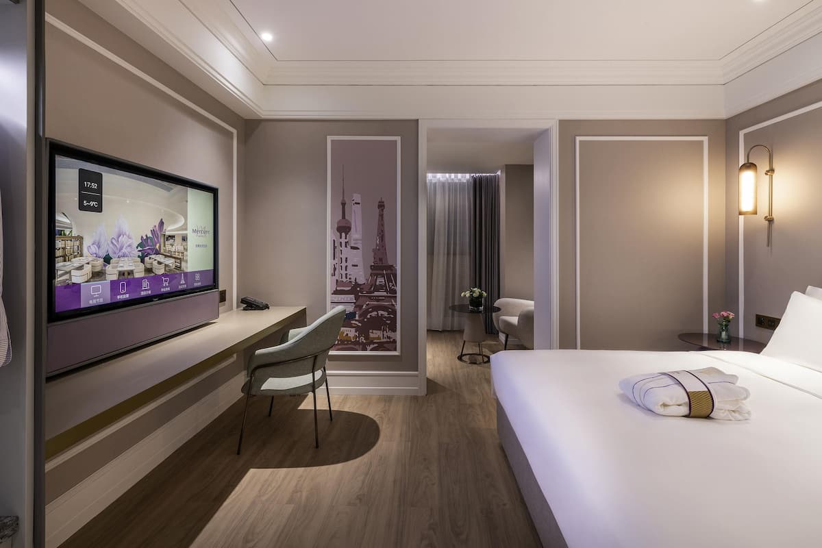 A modern hotel room with a large bed, a desk with a chair, a TV, and a view of a cityscape through a window.
