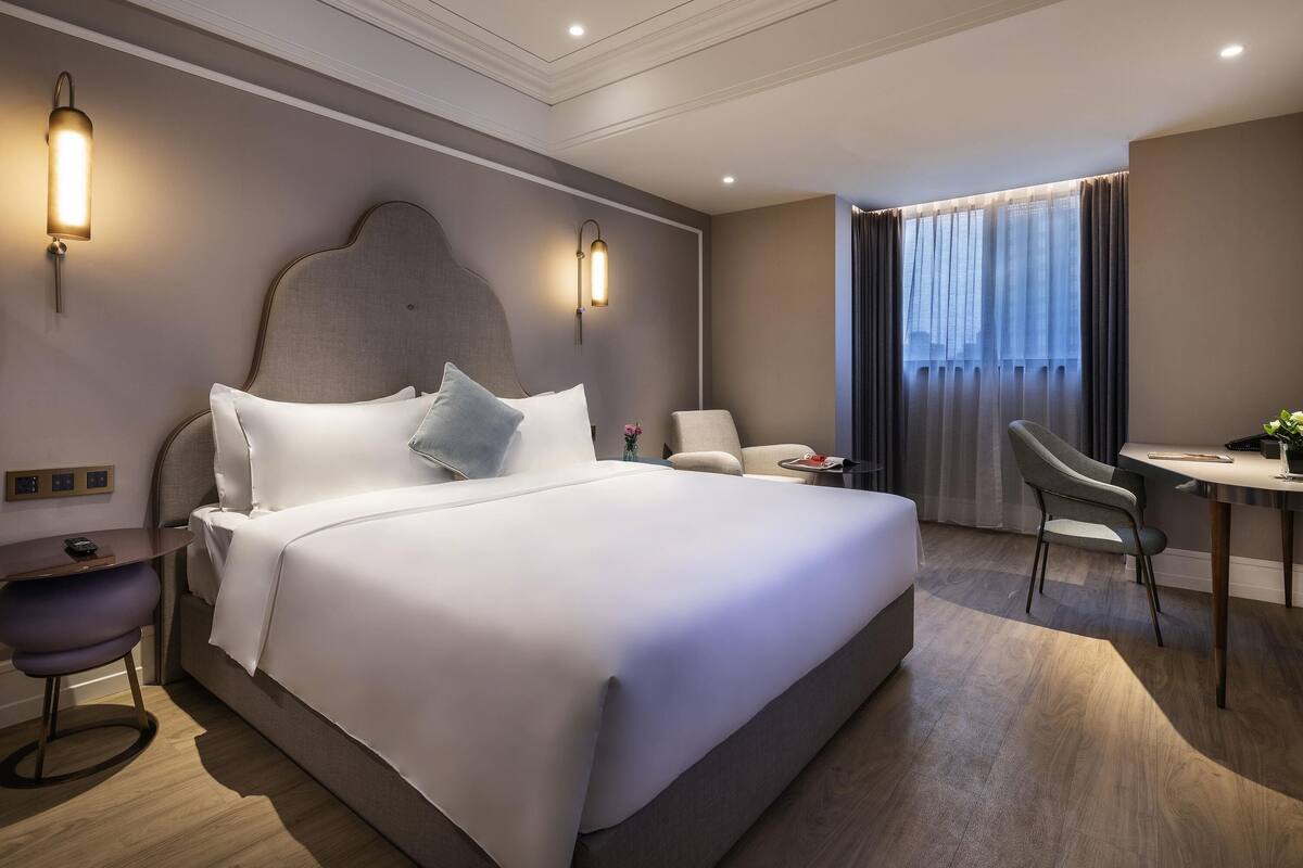 A modern hotel room with a large bed, bedside tables, a desk with a chair, and a view of the city through the window.