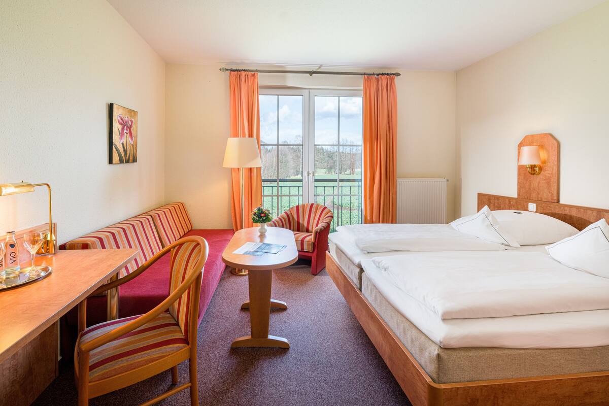 Habitación de hotel con una cama, un escritorio con lámpara, una silla y vistas al exterior a través de una ventana.