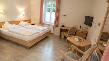 Quarto de hotel com uma cama de casal, uma pequena escrivaninha de madeira com uma cadeira, uma televisão e uma janela com cortinas.