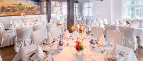 A banquet hall with multiple round tables set for a formal event, featuring white tablecloths, silverware, and candles.